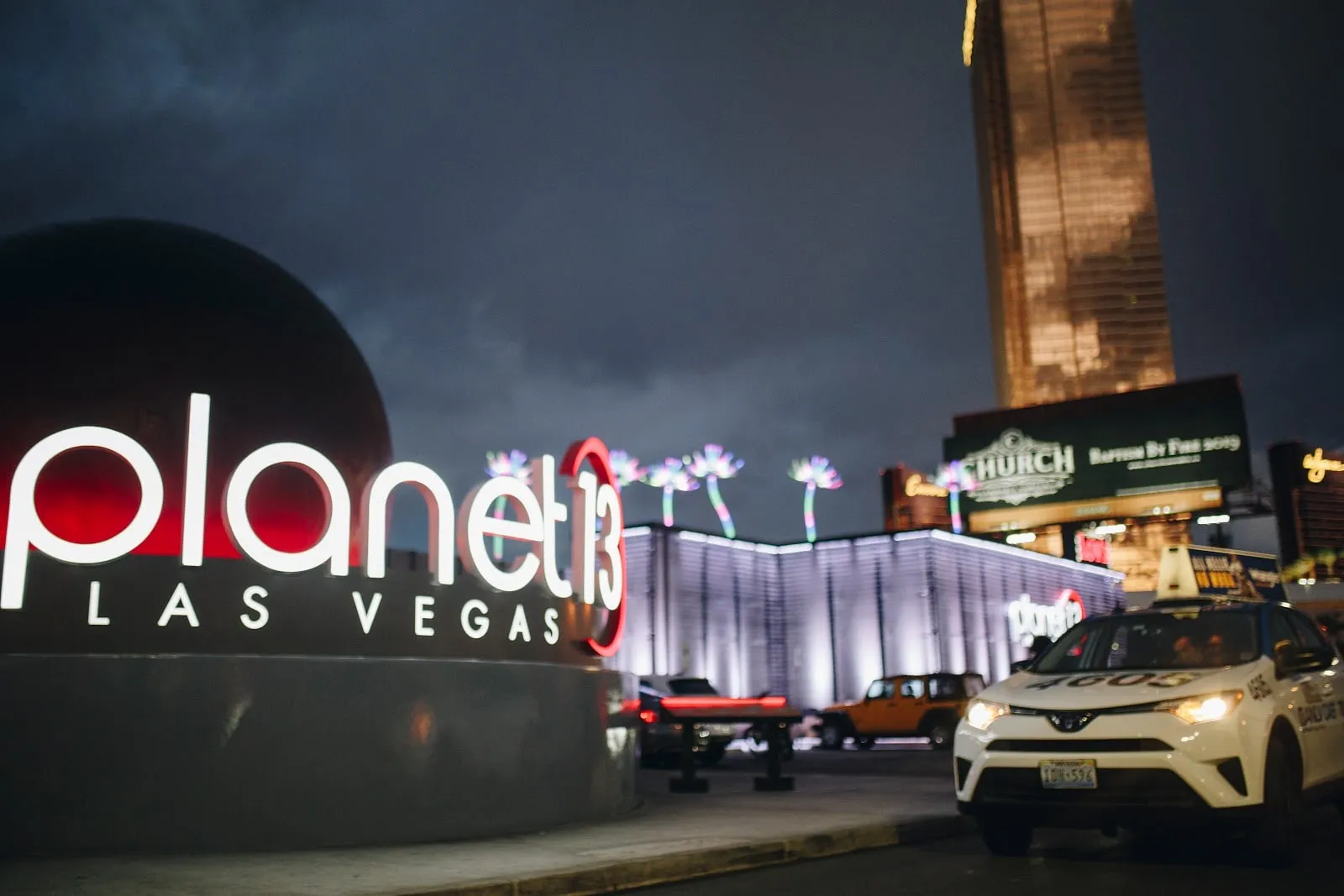 The Planet 13 sign lit at night, a taxi pulling in